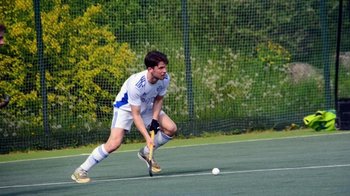 Hockeyspieler in heller Kleidung beim Hockeyspielen auf dem Spielfeld im Freien, mit Bäumen im Hintergrund.