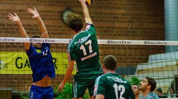 Handballspieler in grünen und blauen Trikots beim Sprungwurf am Netz während eines Hallenhandballspiels.