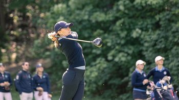 Golfspielerin beim Abschlag auf dem Golfplatz, im Hintergrund stehen weitere Golfer vor einer grünen Baumkulisse.