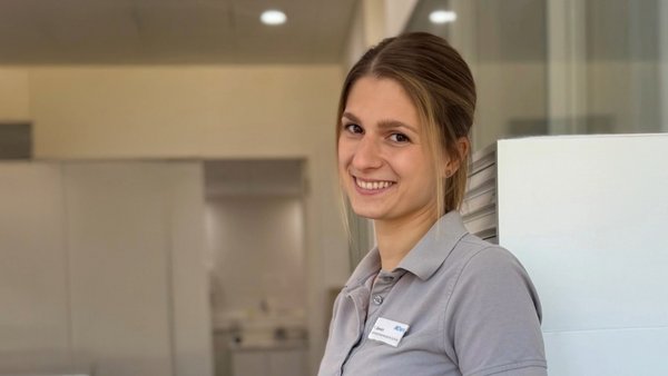 Zahntechnikerin trägt ein graues Shirt, schaut in die Kamera und lächelt