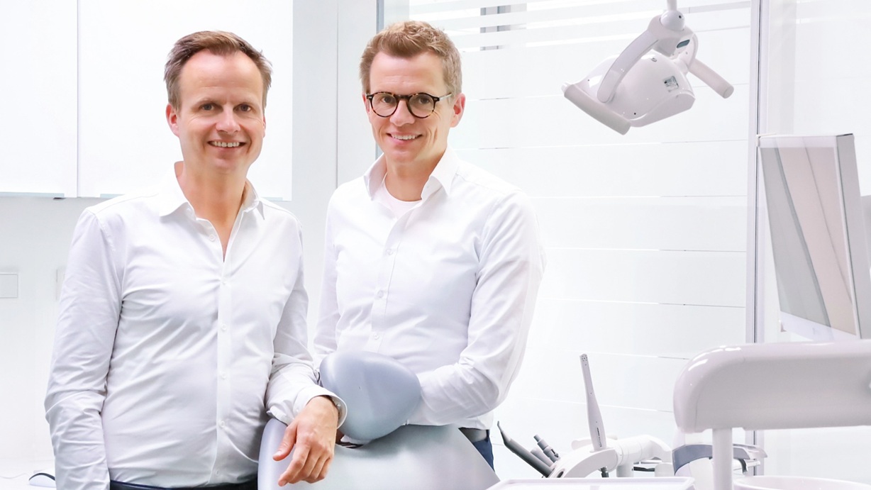 Die Unternehmensgründer lehnen am Behandlungsstuhl und lächeln in die Kamera